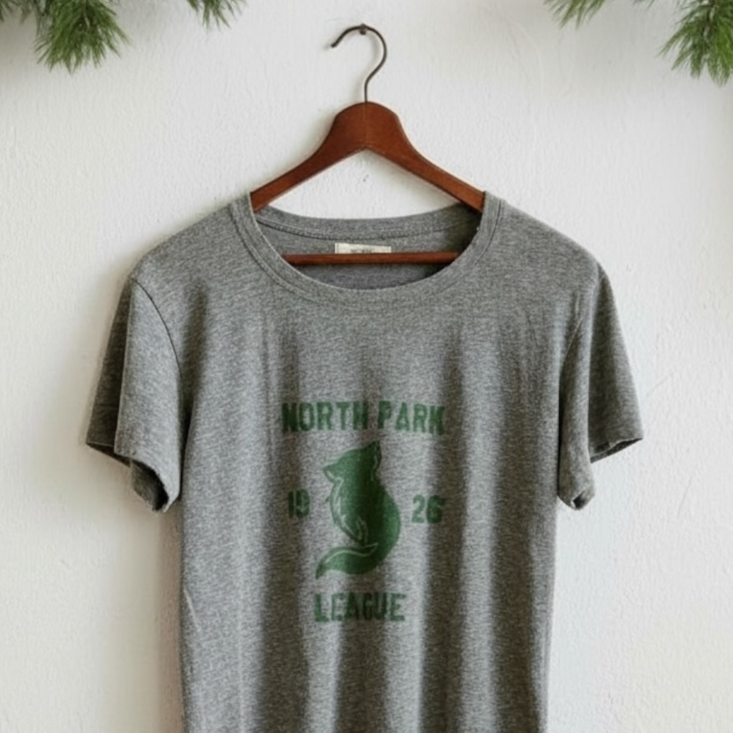 Gray t-shirt with green graphic and text on a wooden hanger against a white wall.